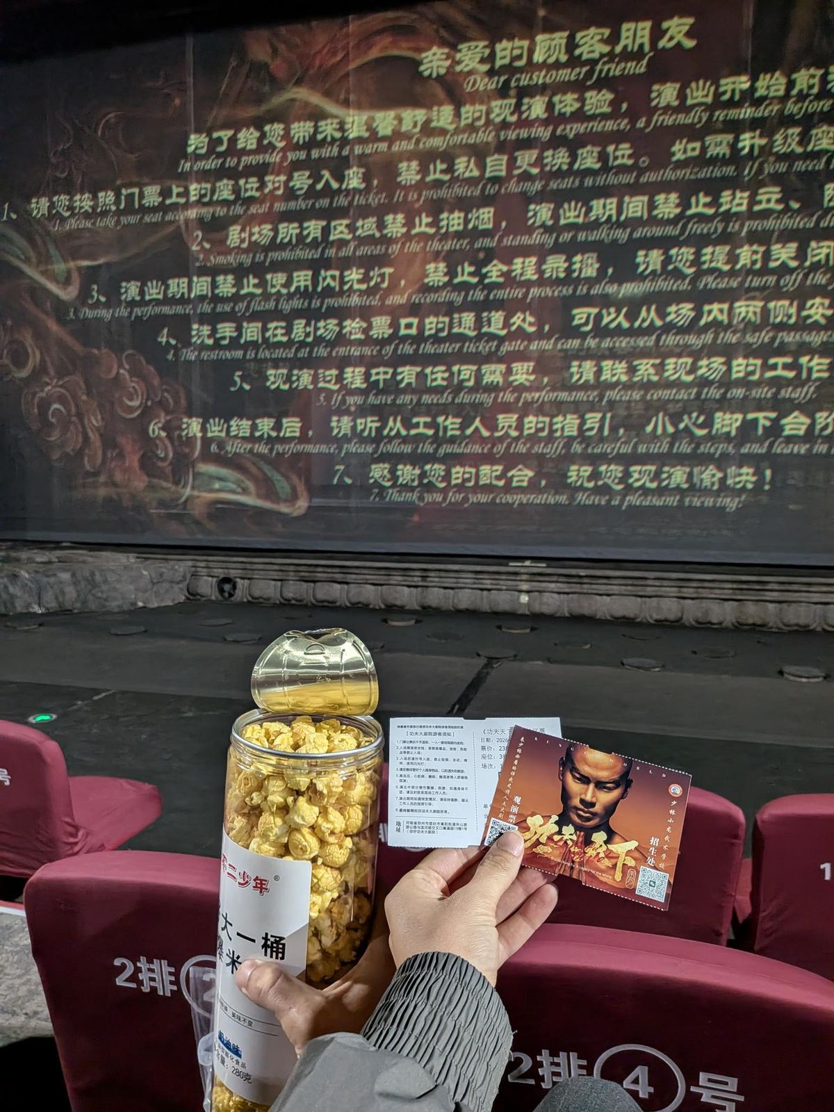 Dans le théâtre, un seau de popcorn 不二少年 et deux billets du show 功夫天下 dans les mains, écran de scène et fauteuils rouges 2排 4号 derrière