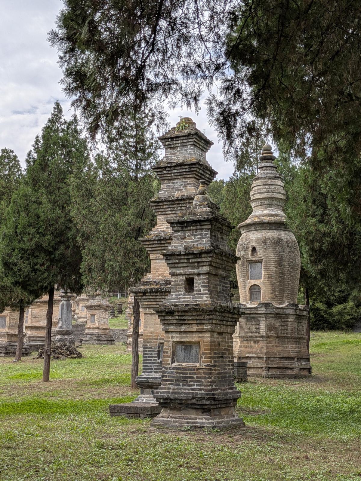 Stupa de brique à toits superposés et stupa à dôme oignon dans une clairière d'herbe, conifères, ciel couvert, stèles en arrière-plan