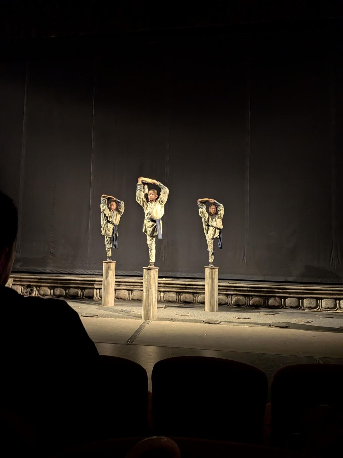 Trois acrobates en costumes clairs équilibrés sur des piliers de bois cylindriques, bras levés, scène noire et bord sculpté, fauteuils d'audience en silhouette au premier plan