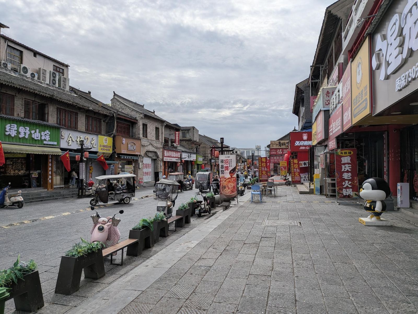 Rue commerçante de la vieille ville, bâtiments en brique de deux étages, enseignes 绿野仙踪, 重庆老火锅, 张亮麻辣烫, panneaux 全场2元 et 19.8元, quelques scooters, ciel couvert