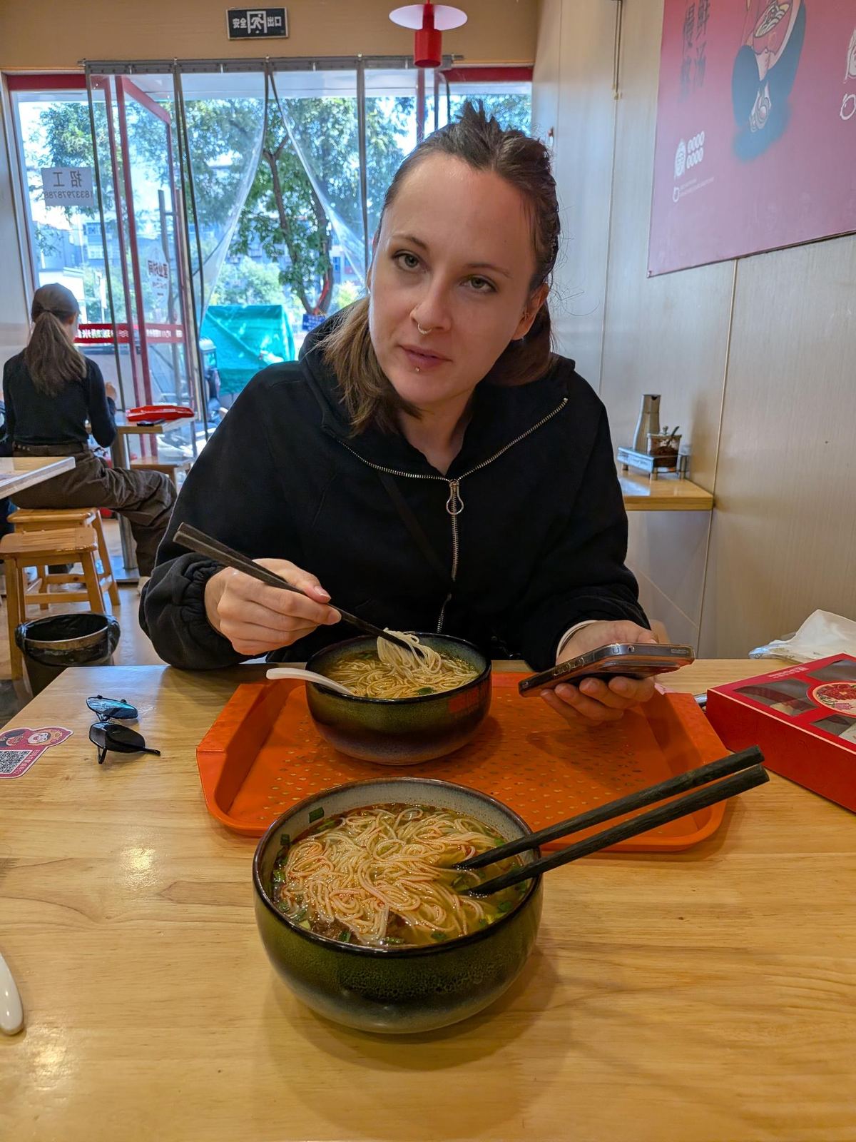 Marion assise dans le petit resto de nouilles, baguettes levées au-dessus de sa gamelle, bol de nouilles sur plateau orange, porte vitrée en arrière-plan