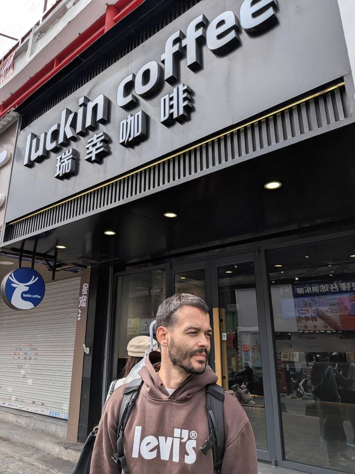 Tristan en hoodie marron et sac à dos devant la vitrine d'un Luckin Coffee, enseigne grise luckin coffee 瑞幸咖啡, logo cerf rond bleu, auvent baissé