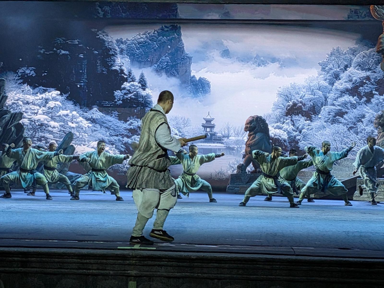 Moine au gilet gris et bâton en posture de combat, troupe de moines en robe bleue clair derrière, décor peint de montagne enneigée, pin givré et pagode au loin