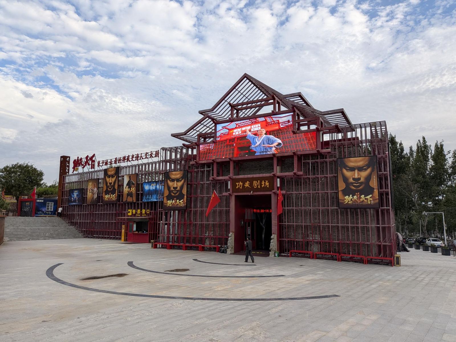 Façade du Kung Fu Theatre en treillis rouge, écran LED géant montrant un martial-artist en robe bleue, affiches 功夫天下, drapeaux et guichet de billetterie, place vide sous un ciel bleu nuageux