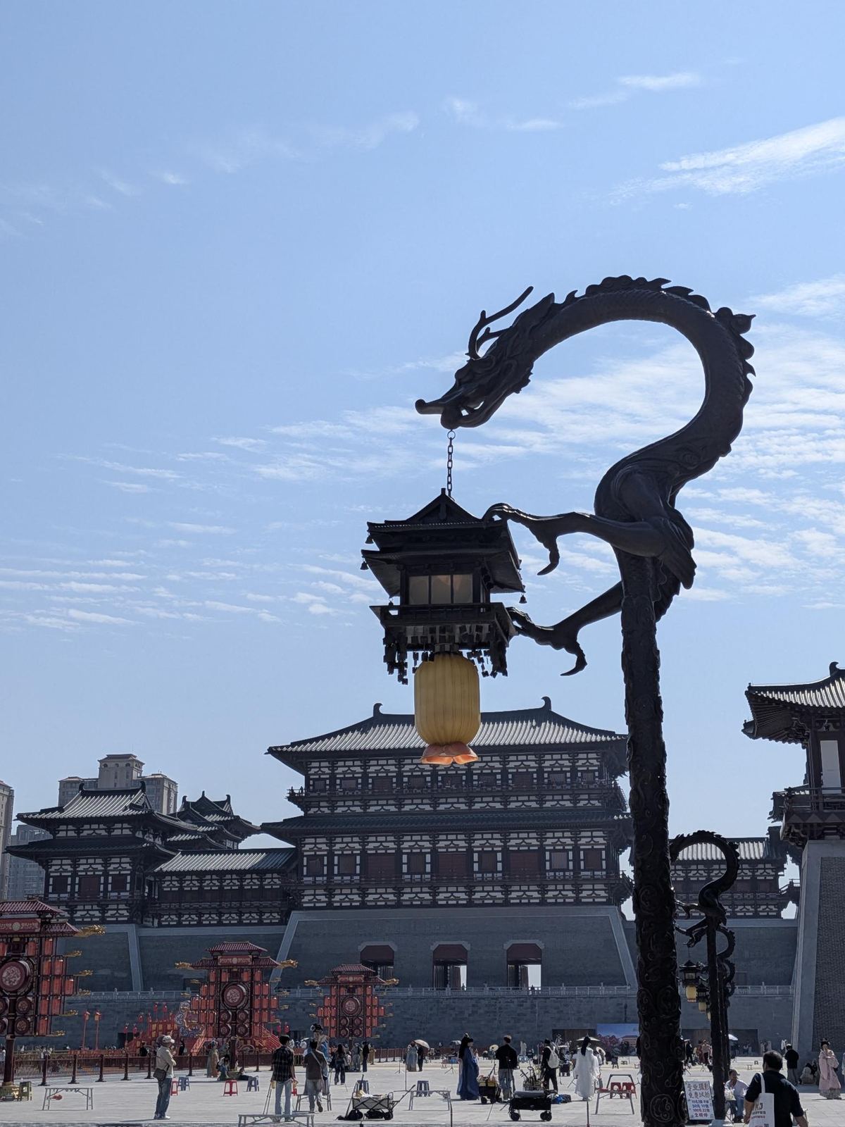 Statue de bronze d'un dragon enroulé dont la gueule suspend une lanterne chinoise lumineuse, palais Tang à plusieurs niveaux derrière, promeneurs en hanfu sur la place