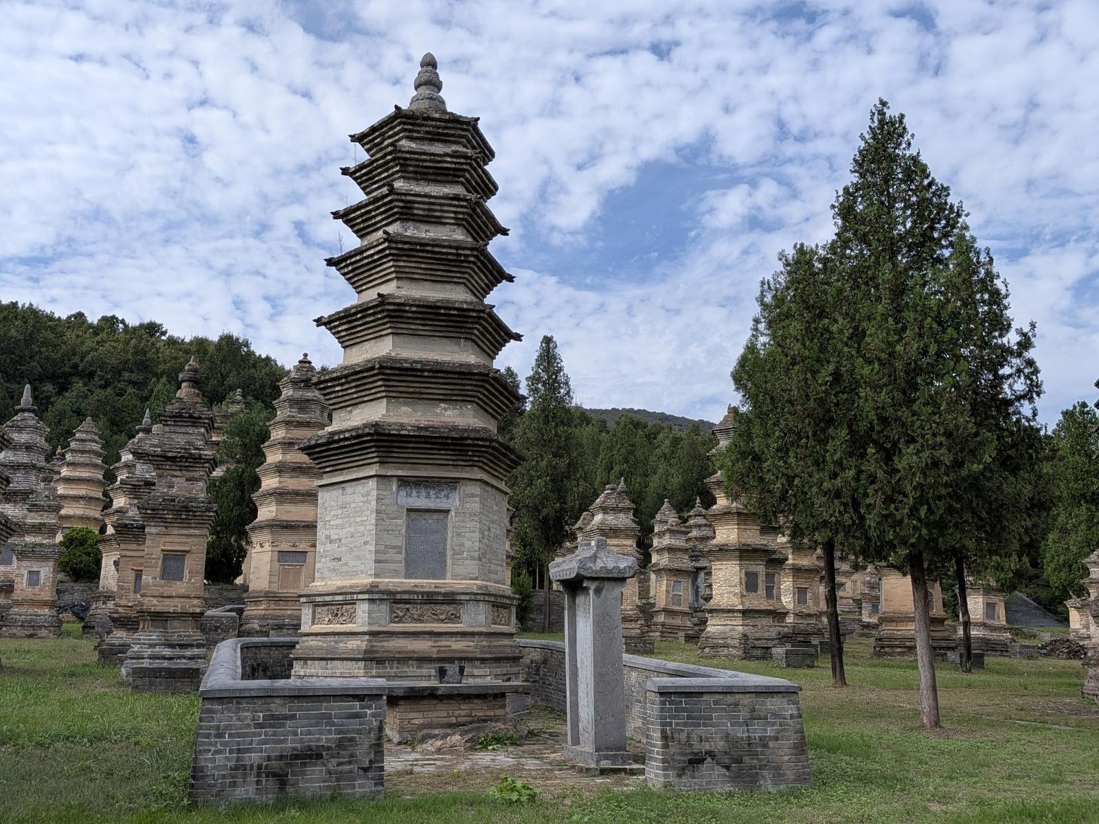 Haute pagode octogonale à toits superposés entourée d'un mur de brique, stèle devant, dizaines de stupa plus petits sur la pelouse, cypres et collines boisées au loin