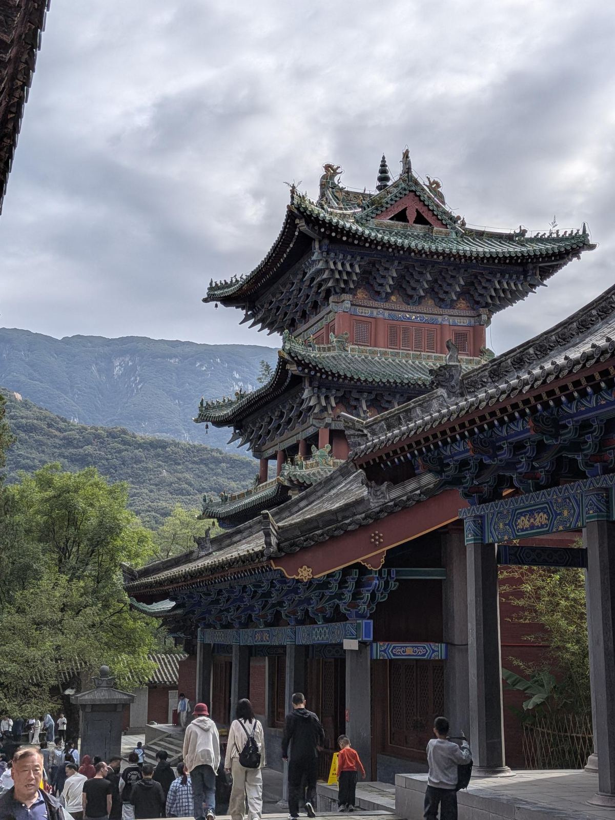 Le complexe du temple au pied de la montagne, pavillon à toits verts et colonnes rouges, foule dans la cour, ciel gris