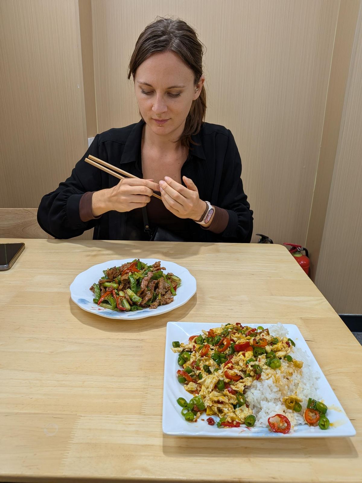 Marion en chemise noire devant sa table, baguettes levées, assiette de lanières de viande et plat d'œufs au piment sur le riz