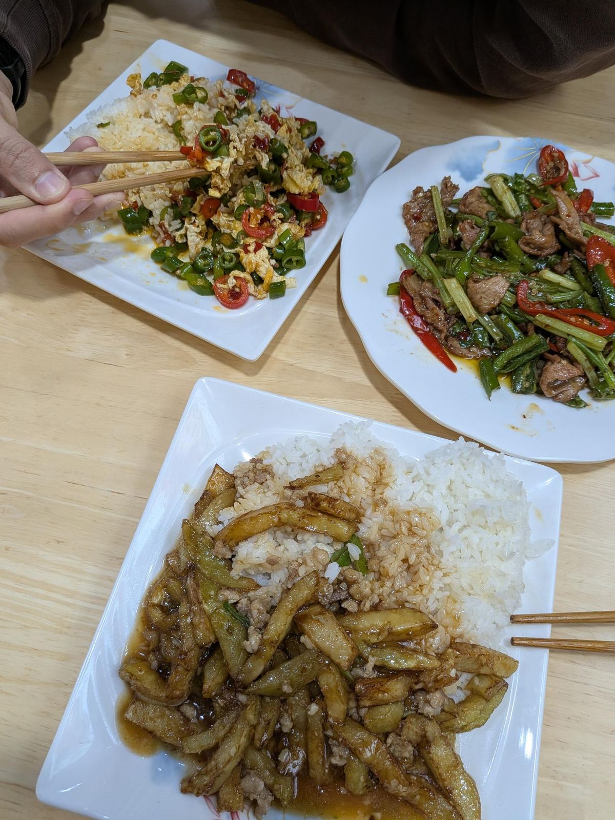 Trois plats sur table en bois clair : riz et œufs brouillés sautés au piment vert et rouge, lanières de viande sautées aux tiges d'oignon, aubergines braisées à la viande hachée, baguettes en travers