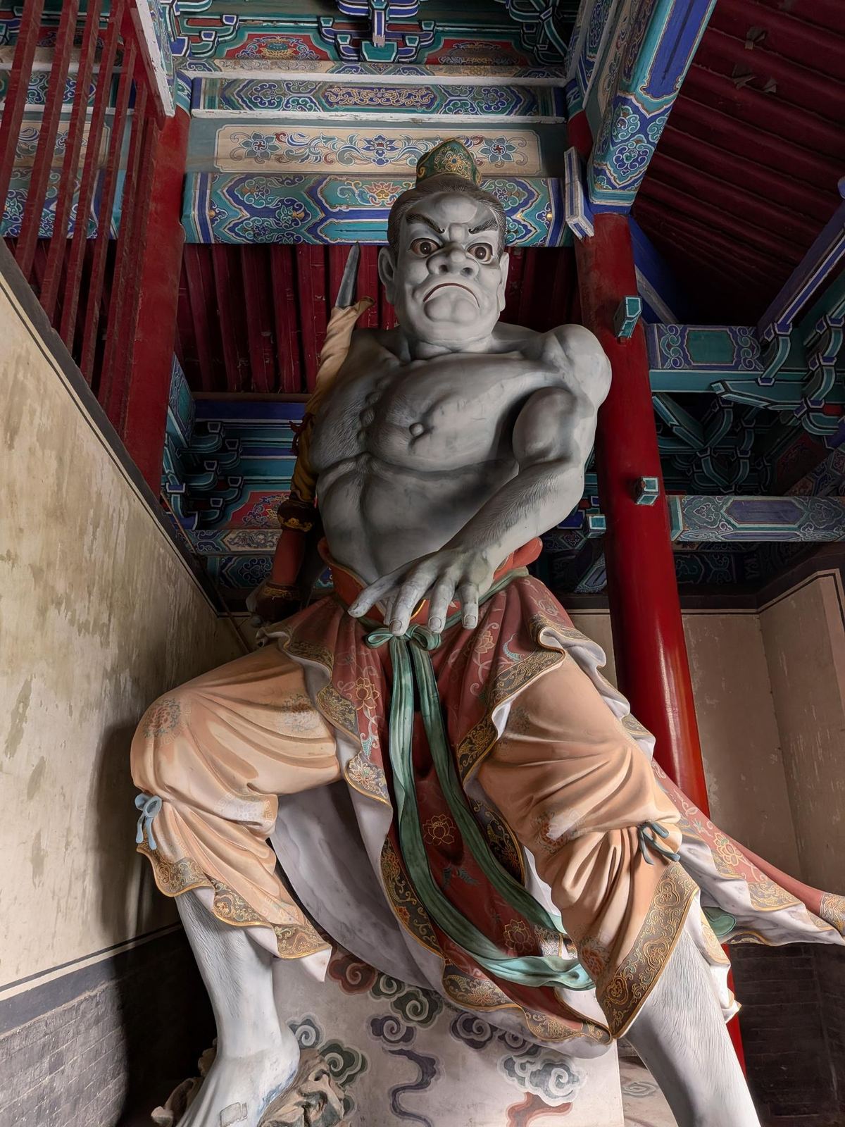 Statue d'un gardien au torse musclé, robe pêche et rouge à liseré doré, sangle verte, devant les encorbellements bleus et dorés du hall