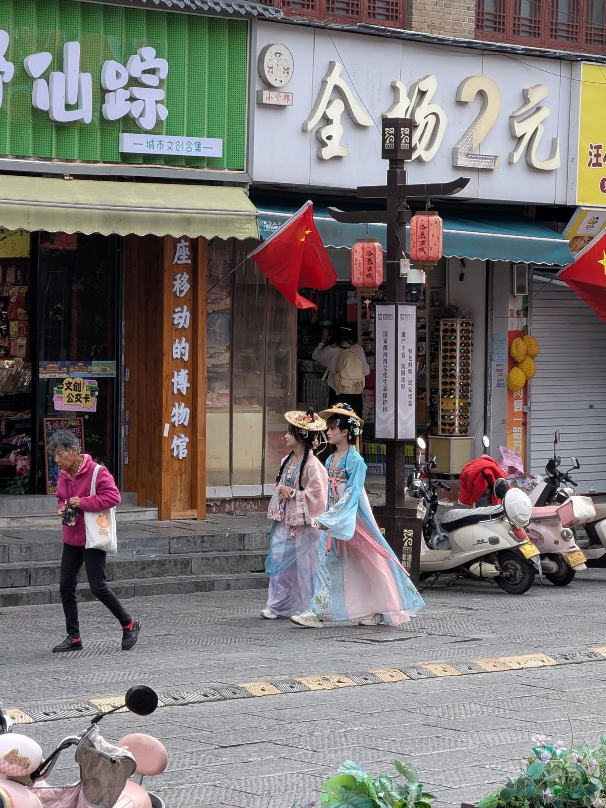 Deux jeunes femmes en hanfu et chapeaux de paille sous les lanternes rouges de 洛邑古城, enseignes 仙踪 et 全场2元, rue pavée et scooters stationnés